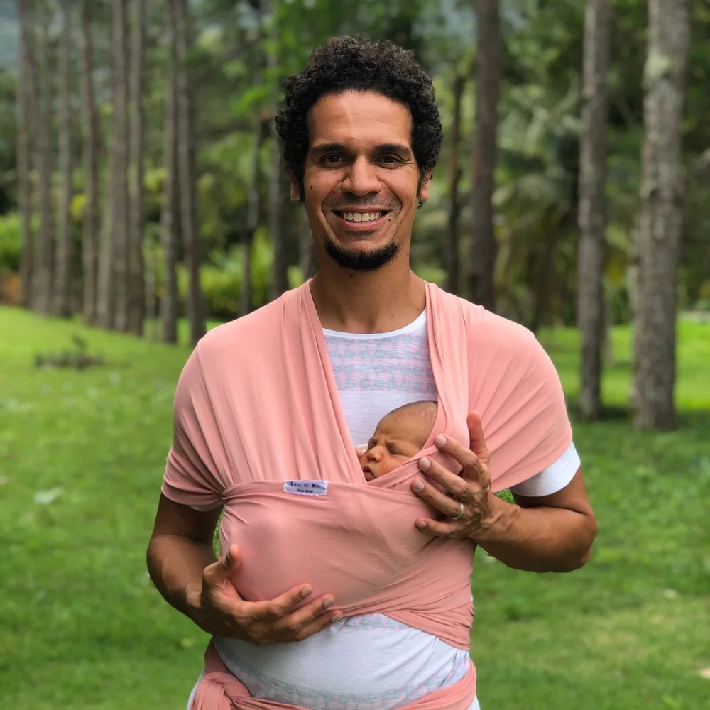 Elcio Leite sorrindo, com a filha recém-nascida aconchegada num sling